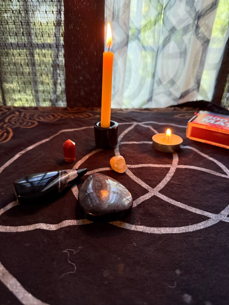 A lit yellow candle and a small candle sit on a dark cloth with a geometric design. Various stones, including crystals, are arranged around them, creating a peaceful setup for a ritual.