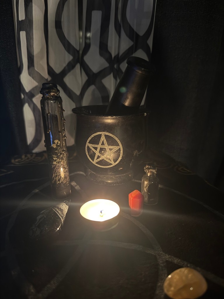 A beautifully arranged witchcraft altar featuring a black cauldron with a pentagram symbol, a lit candle, a red crystal, small glass vials, and various mystical tools set against a dark backdrop.