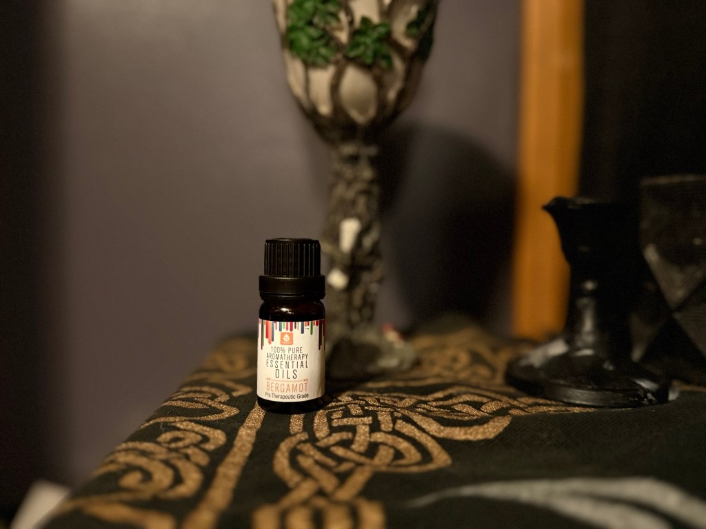 A bottle of bergamot essential oil placed on a patterned altar cloth, alongside a decorative chalice and a black candlestick.