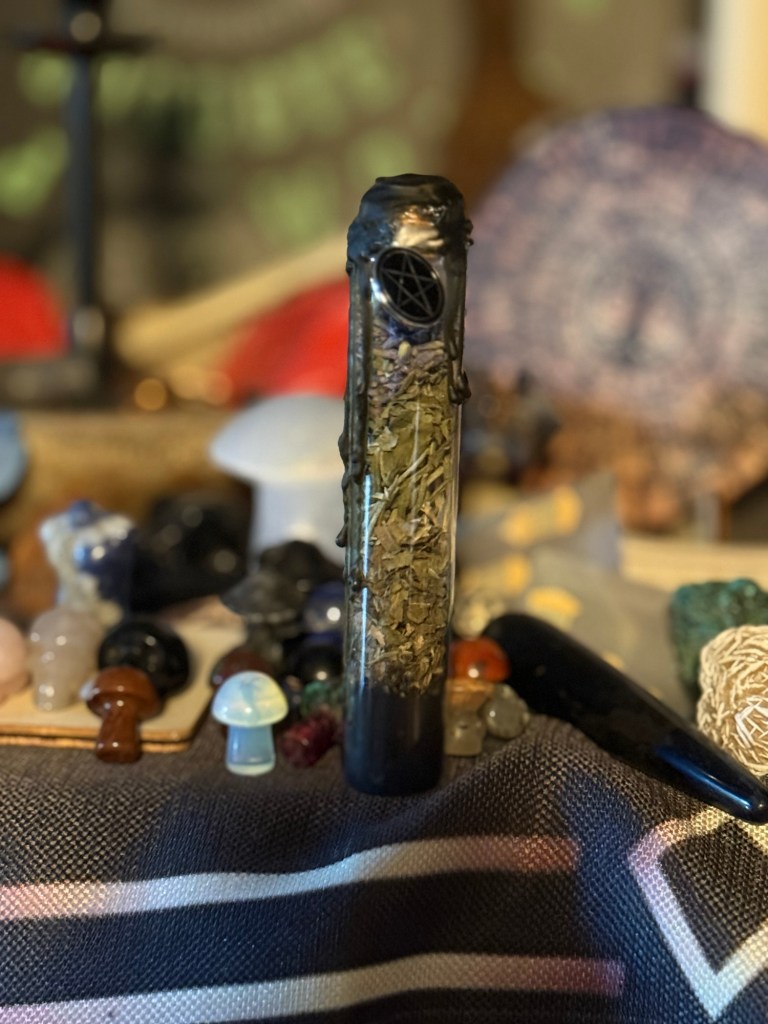 A glass vial filled with herbs, surrounded by various crystals such as mushrooms and skulls on a black sheet. 